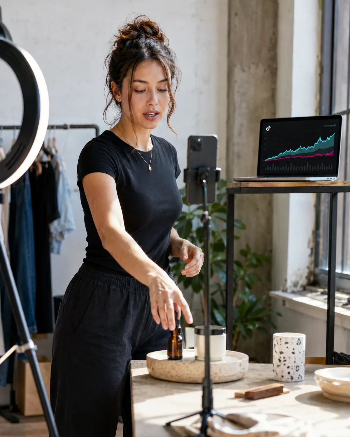 Creator filming a product video on a phone with a TikTok analytics dashboard on the laptop behind her